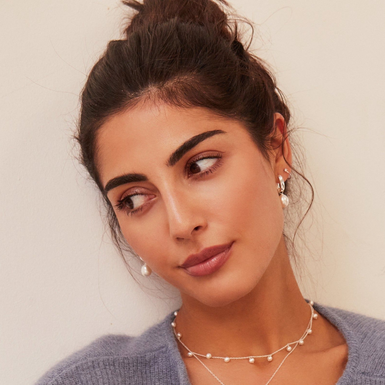 A brunette woman with her hair up wearing silver thick squared hoop pearl drop earrings in her ear lobes and a silver drop pearl choker around her neck