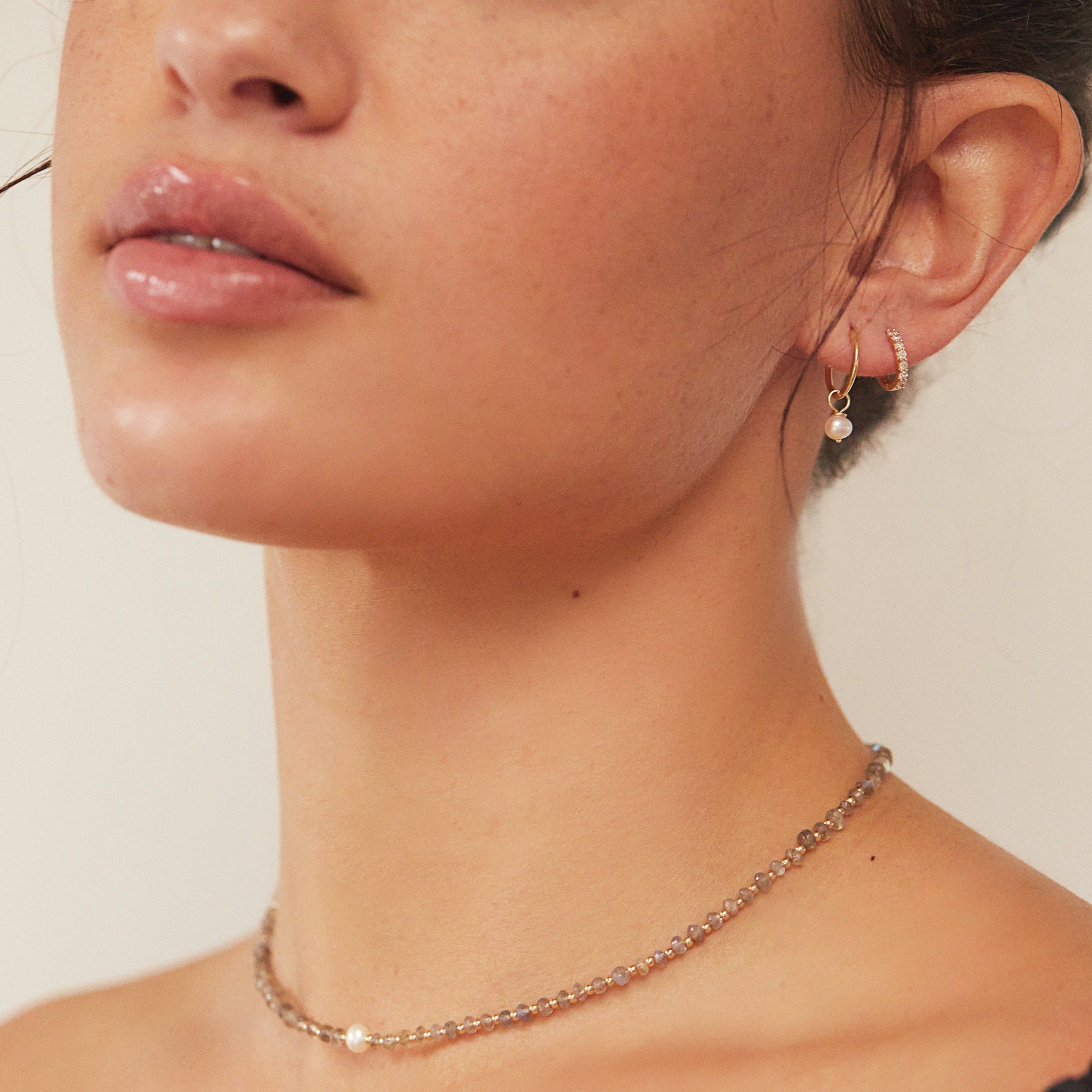 A woman wearing a gold small pearl drop hoop earring and a gold diamond style huggie hoop earring in her ear lobe and a pearl choker around her neck