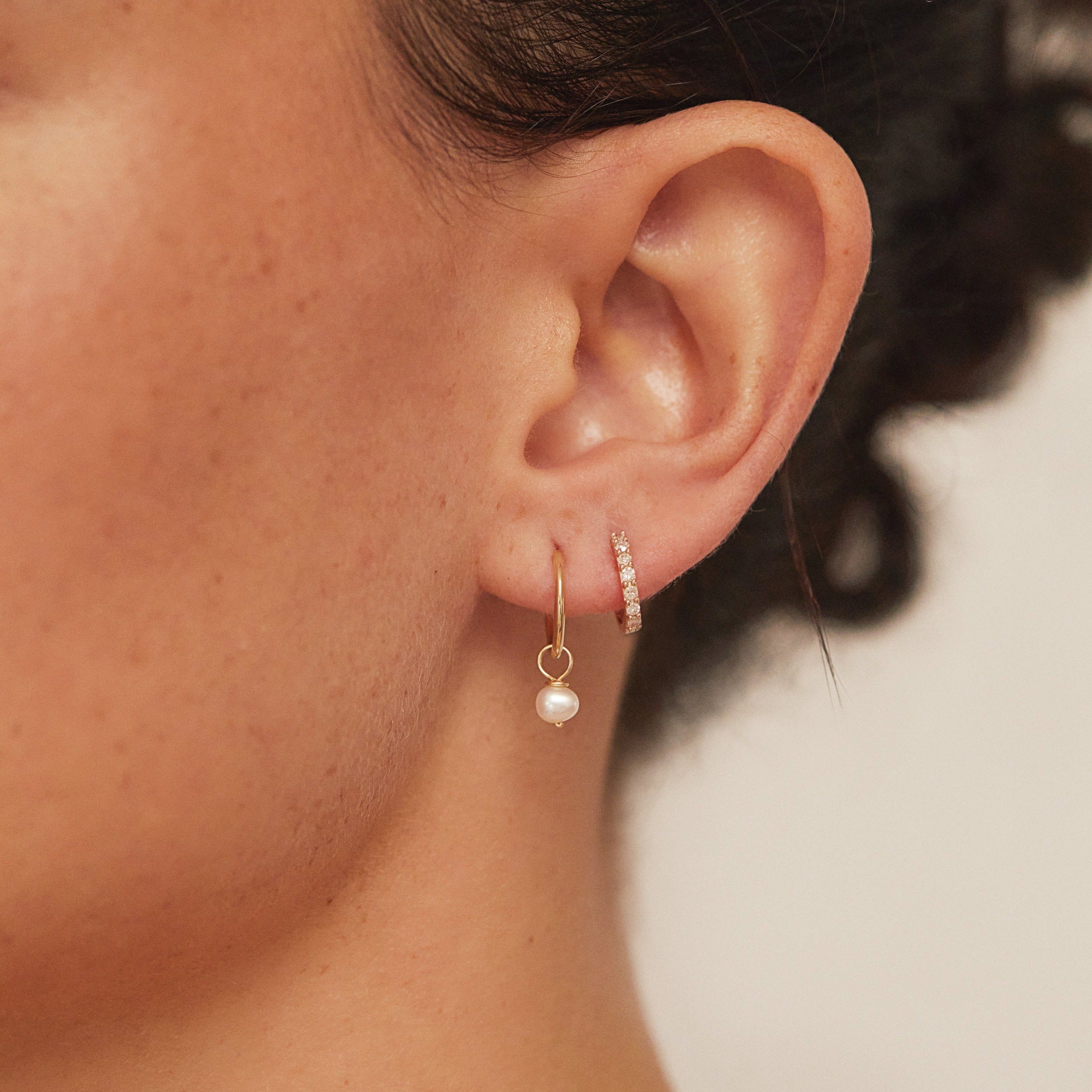 A gold small pearl drop hoop earring and a gold diamond style huggie hoop earring in an ear lobe