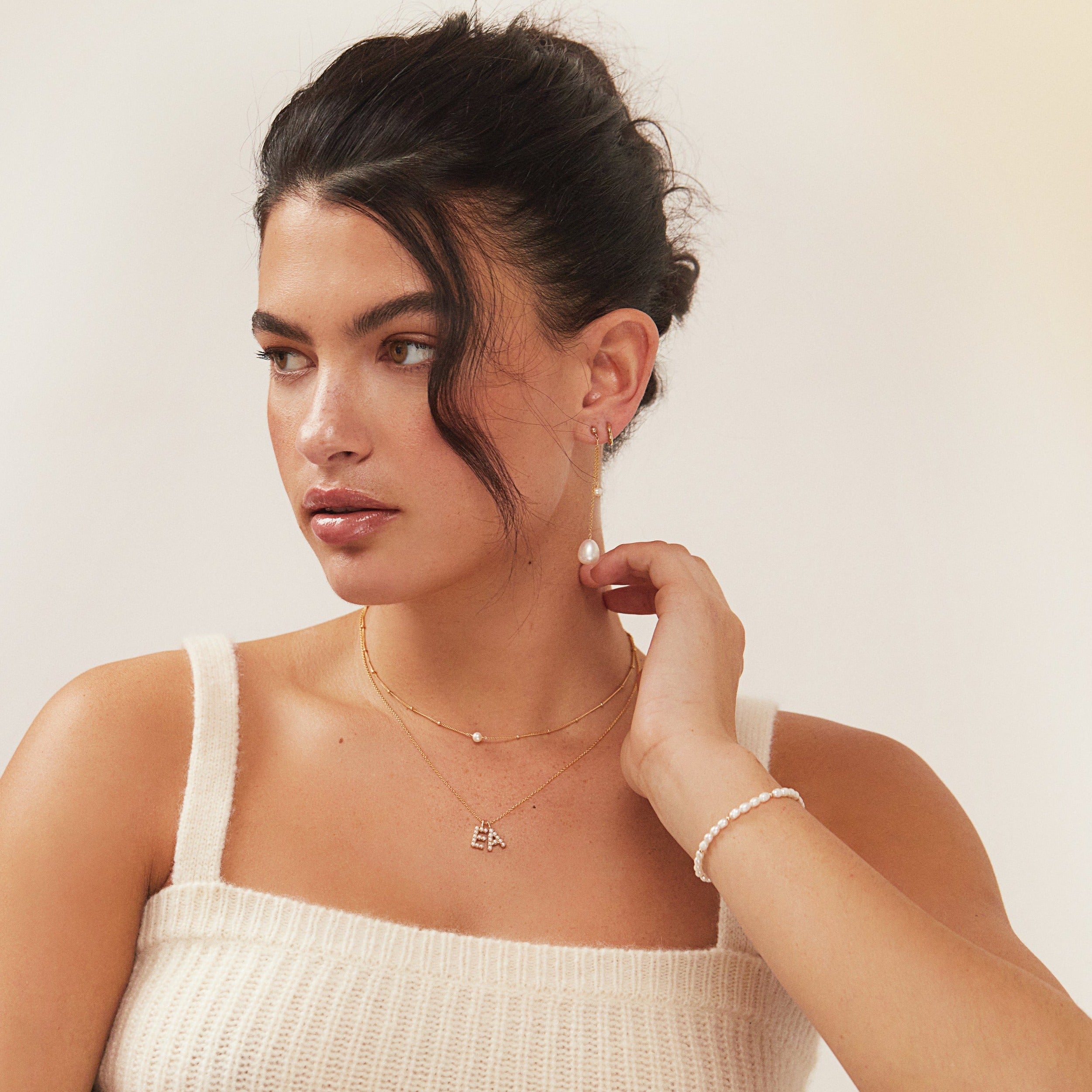 A brunette woman wearing a gold beaded seed pearl bracelet around her wrist and a E and A letter necklace and a gold satellite single pearl choker around her neck