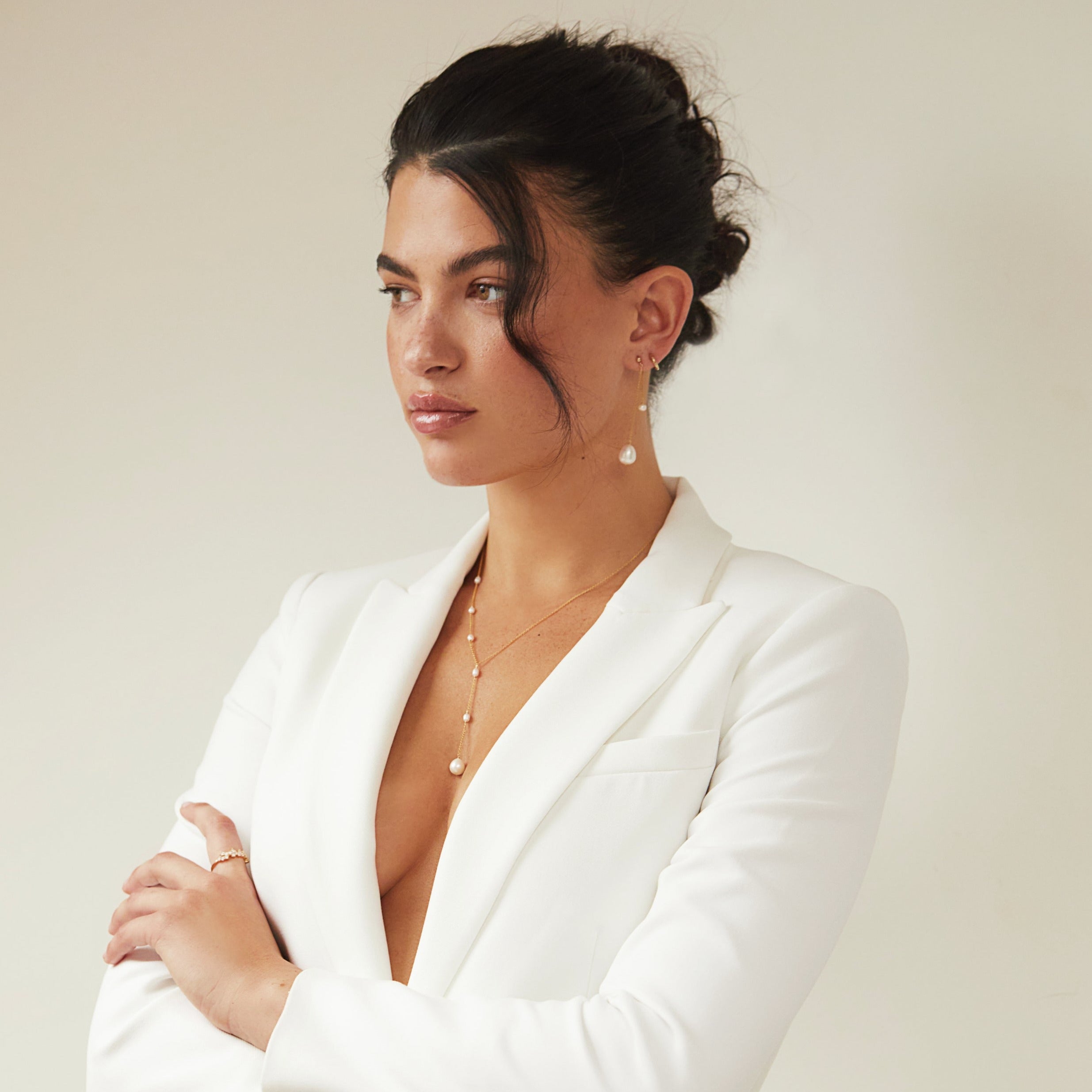Gold seed pearl lariat necklace around a neck of a woman wearing a white blazer and a gold large pearl drop ear threader in her ear lobe, with her arms crossed