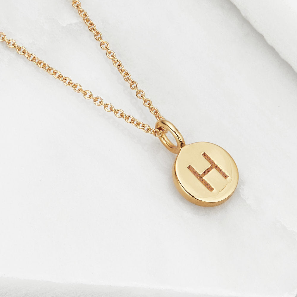 Gold small personalised disc necklace with the letter 'H' engraved on a marble surface