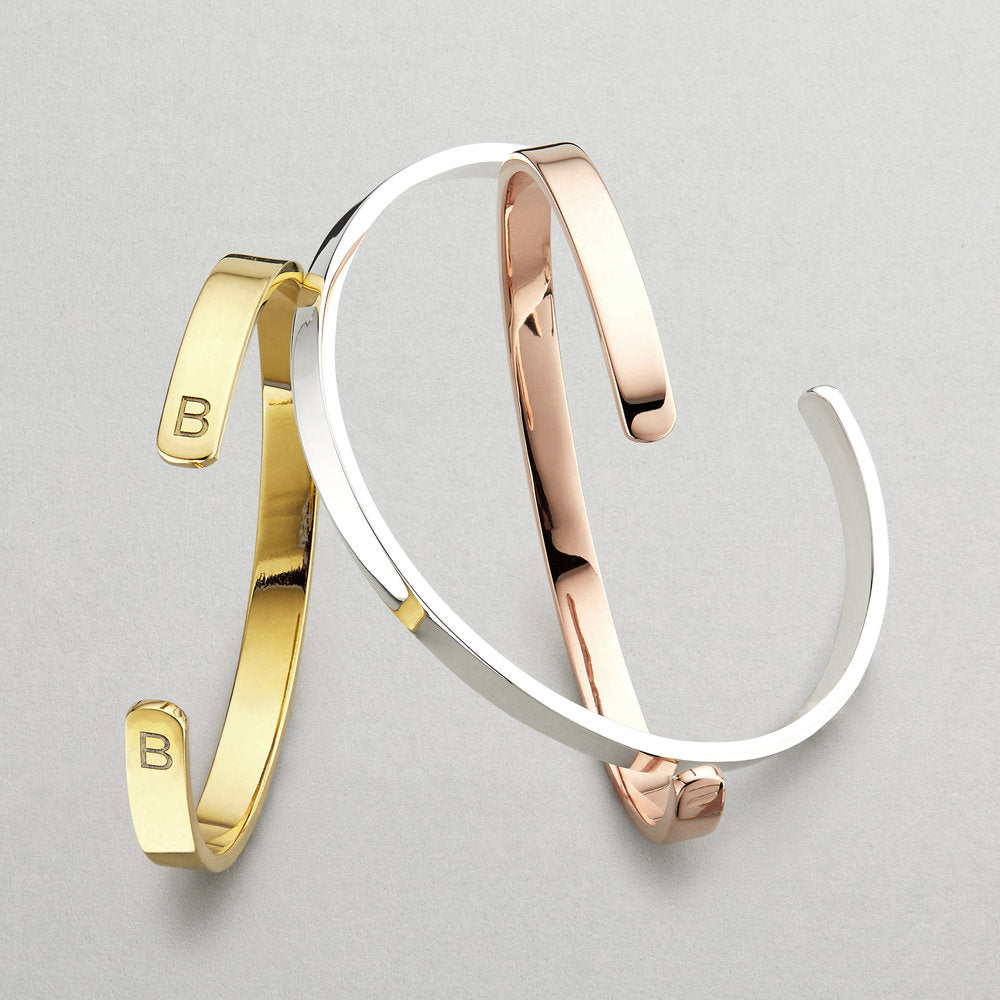 Gold thick engraved bangle with two of the letter 'B' engraved on a grey surface alongside a silver thick engraved bangle and rose gold thick engraved bangle