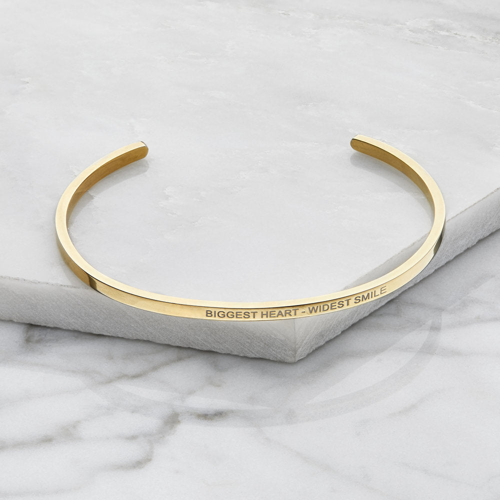 Gold thin engraved bangle engraved with the words 'BIGGEST HEART - WIDEST SMILE' on it, on marble surfaces