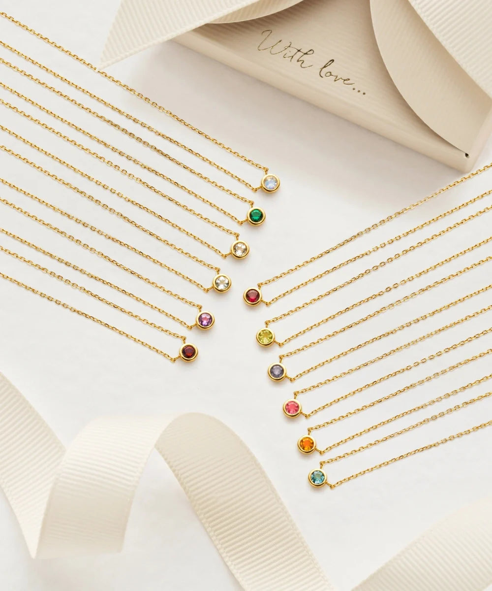 Gold birthstone necklaces arranged with colourful gemstone pendants on a cream background with ribbon detailing.