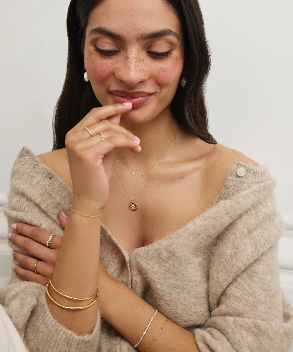 Model wearing layered gold necklaces, pearl earrings, delicate rings and gold bracelets with a neutral knit cardigan.