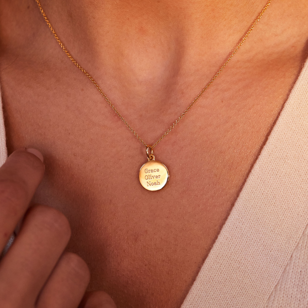 A gold small round engraved disc necklace with the names 'Grace' 'Oliver' and 'Noah' engraved, close up on a woman's chest