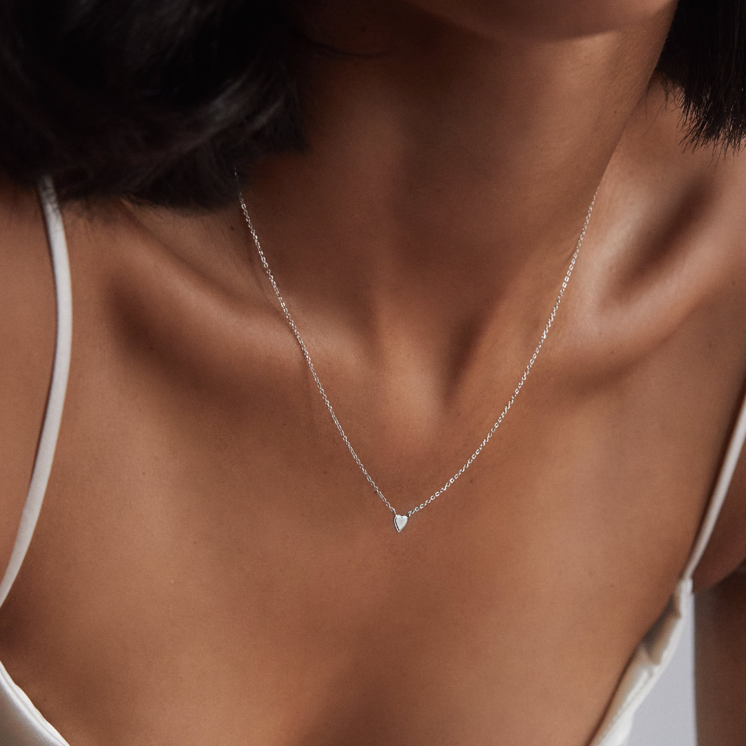 Silver tiny heart necklace around the neck of a woman wearing a white strappy top 