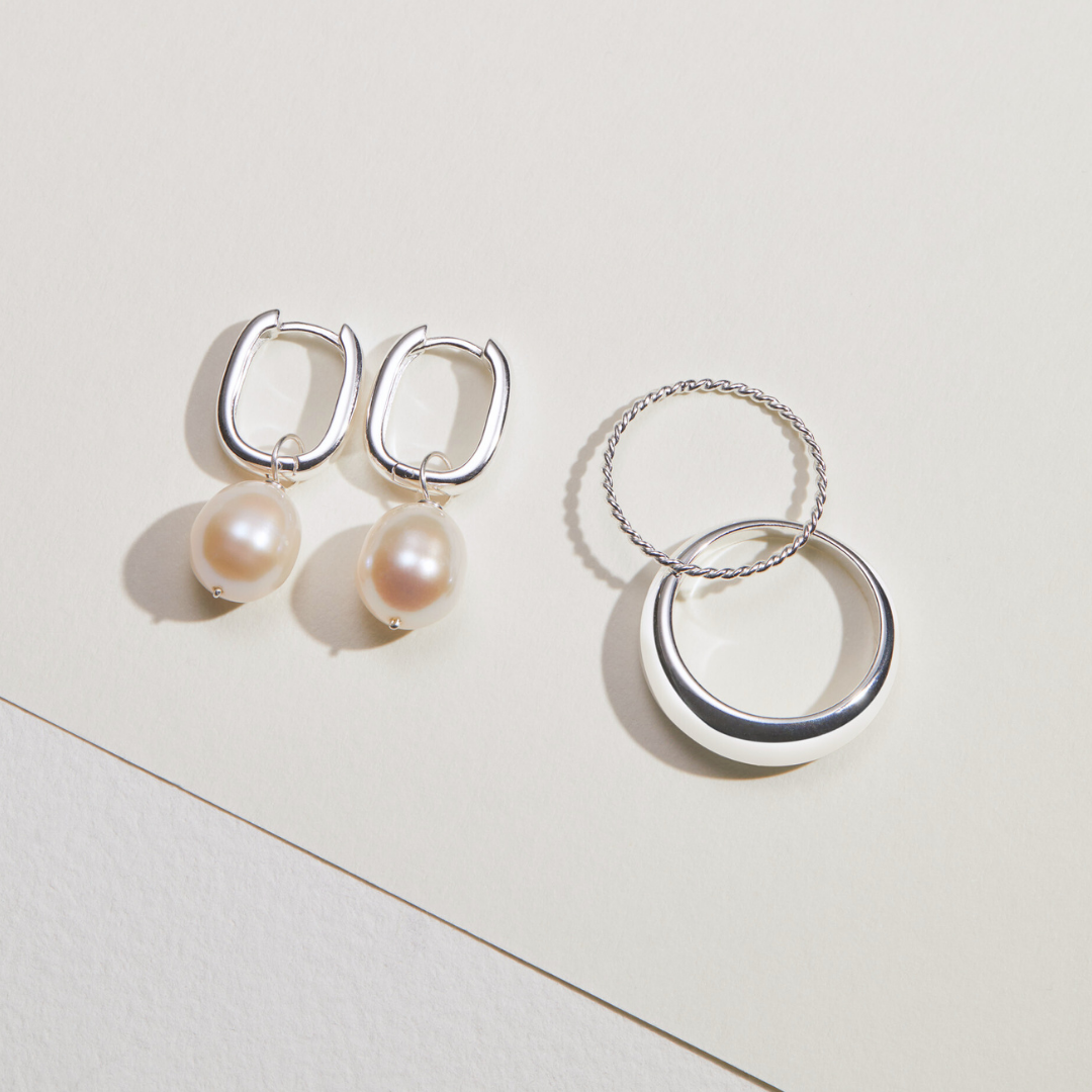 Silver thick squared hoop pearl drop earrings on a paper surface next to silver thin twisted stacking ring and a silver plain dome ring 