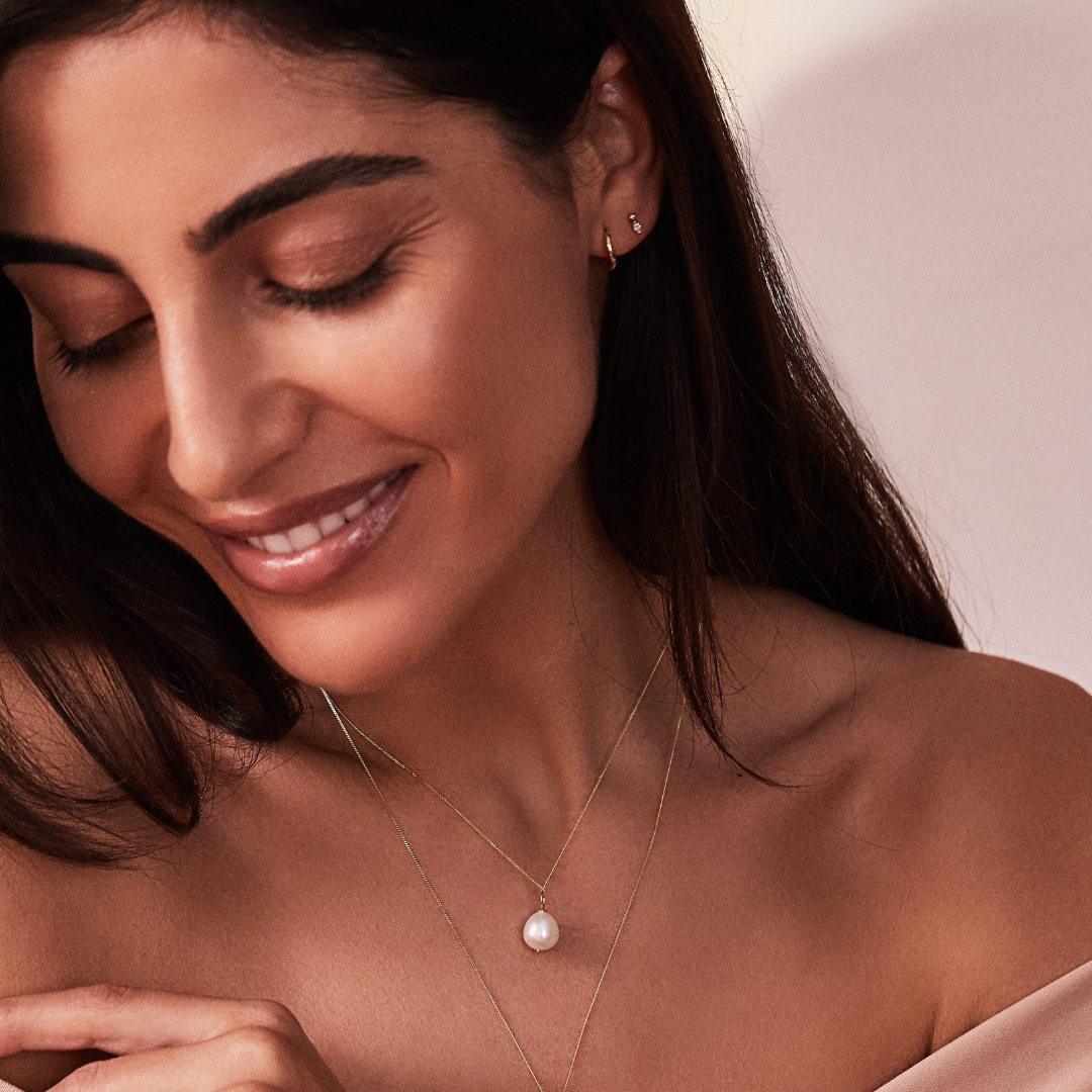A smiling woman looking down wearing a gold wave huggie hoop earring and a stud in one ear lobe and a gold large single pearl necklace around her neck