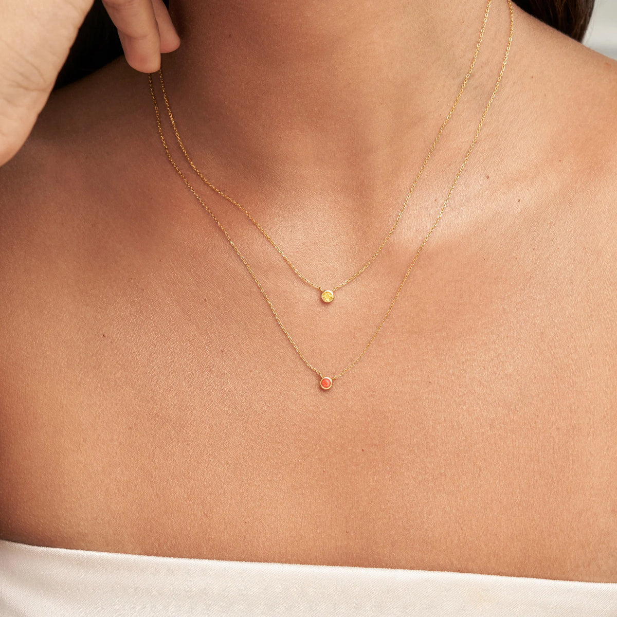 A close-up of a model's neck and upper chest wearing two delicate gold necklaces with small circular pendants, one gold and one coral-pink, styled with a white off-the-shoulder top.