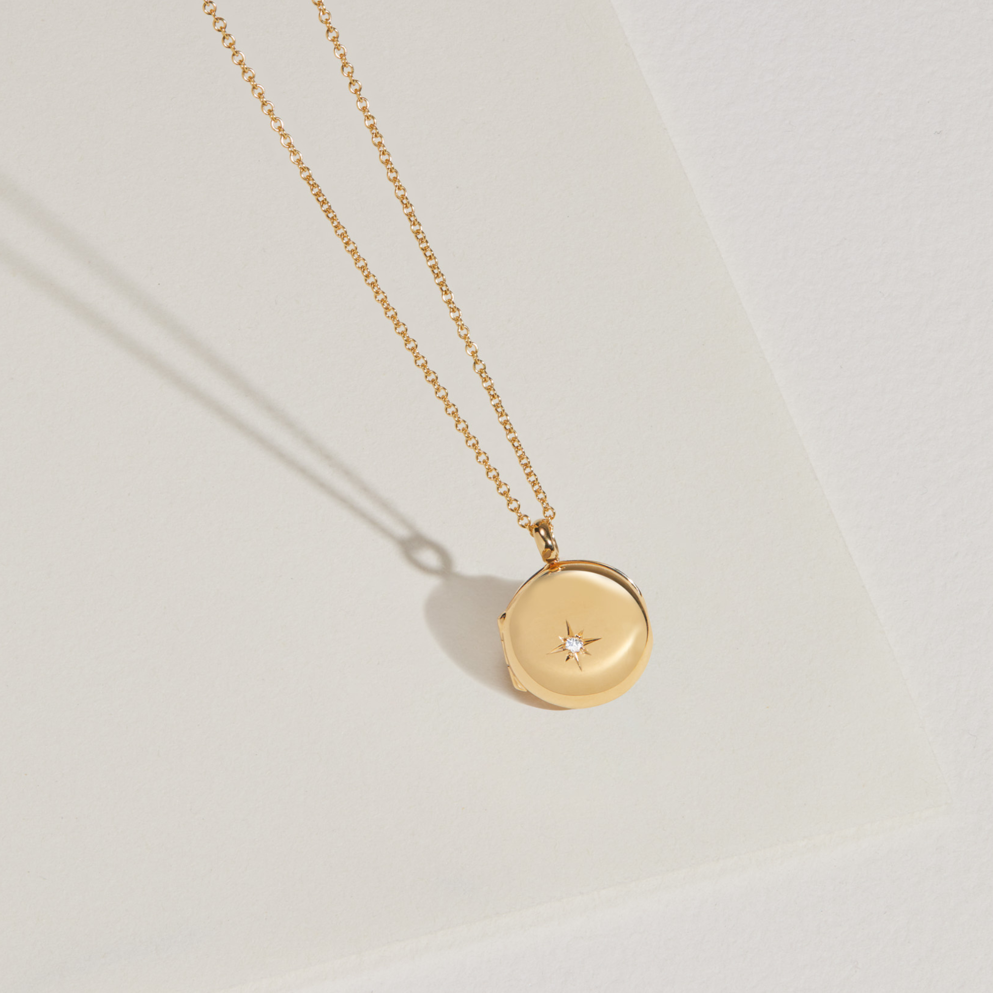 Gold small round diamond locket necklace hanging infront of a paper surface