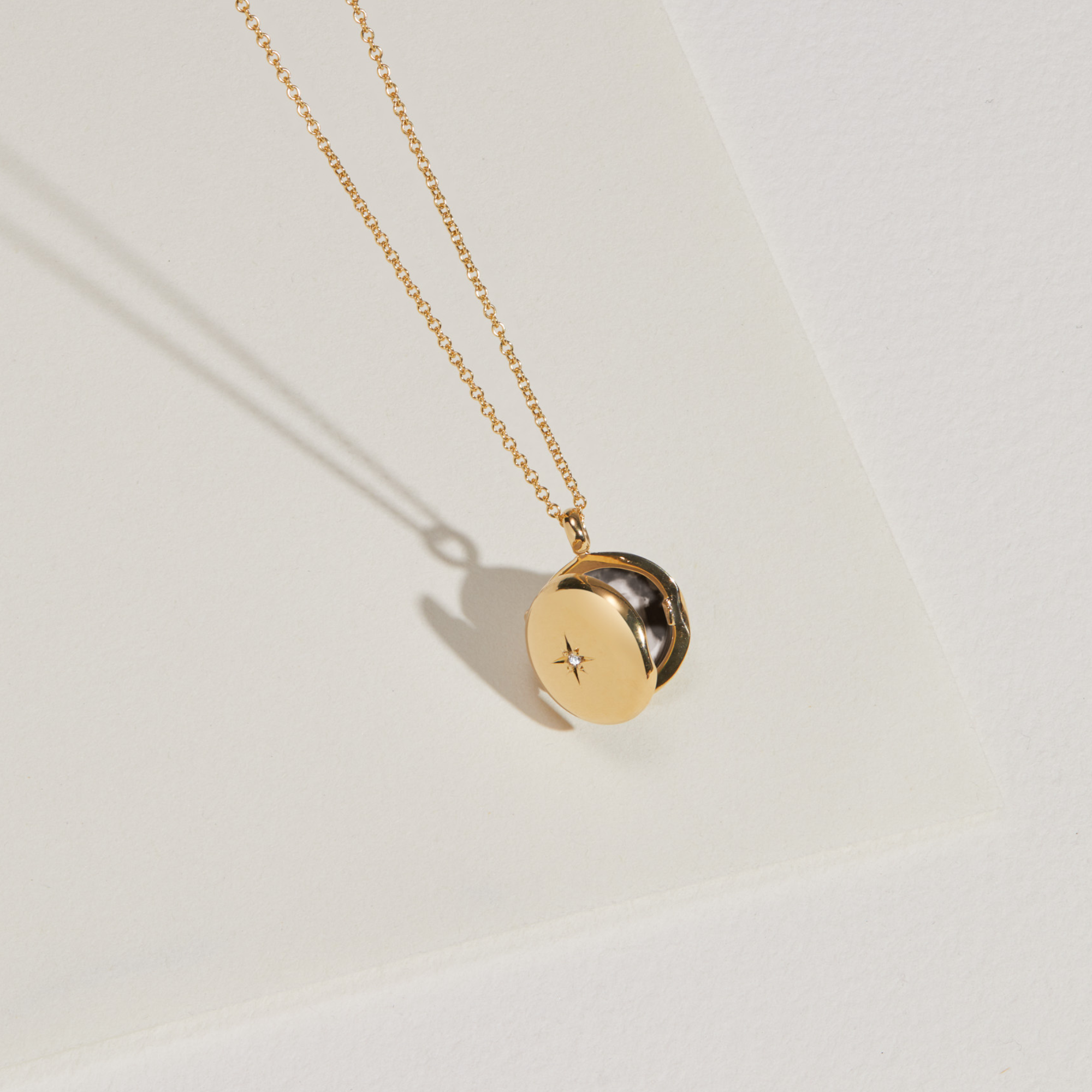Open gold small round diamond locket necklace hanging infront of a paper surface