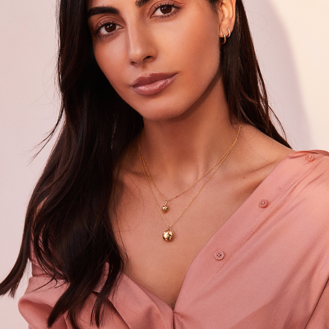 Gold heart pendant necklace around a brunette woman's neck layered with a gold small round diamond locket necklace and wearing a pink shirt
