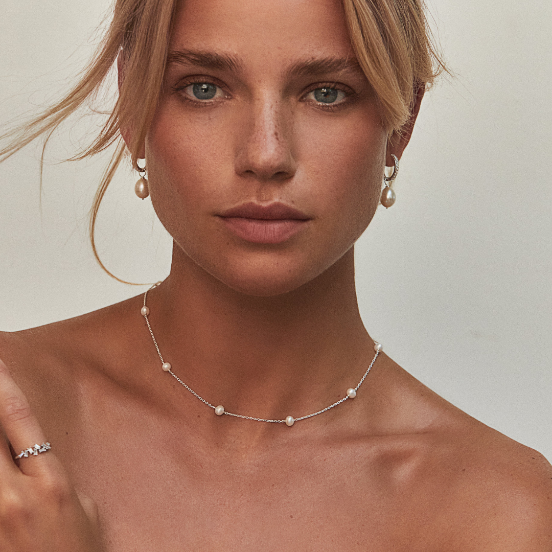 A blonde woman wearing silver diamond style large pearl drop hoop earrings in her ear lobes and a silver pearl choker around her neck