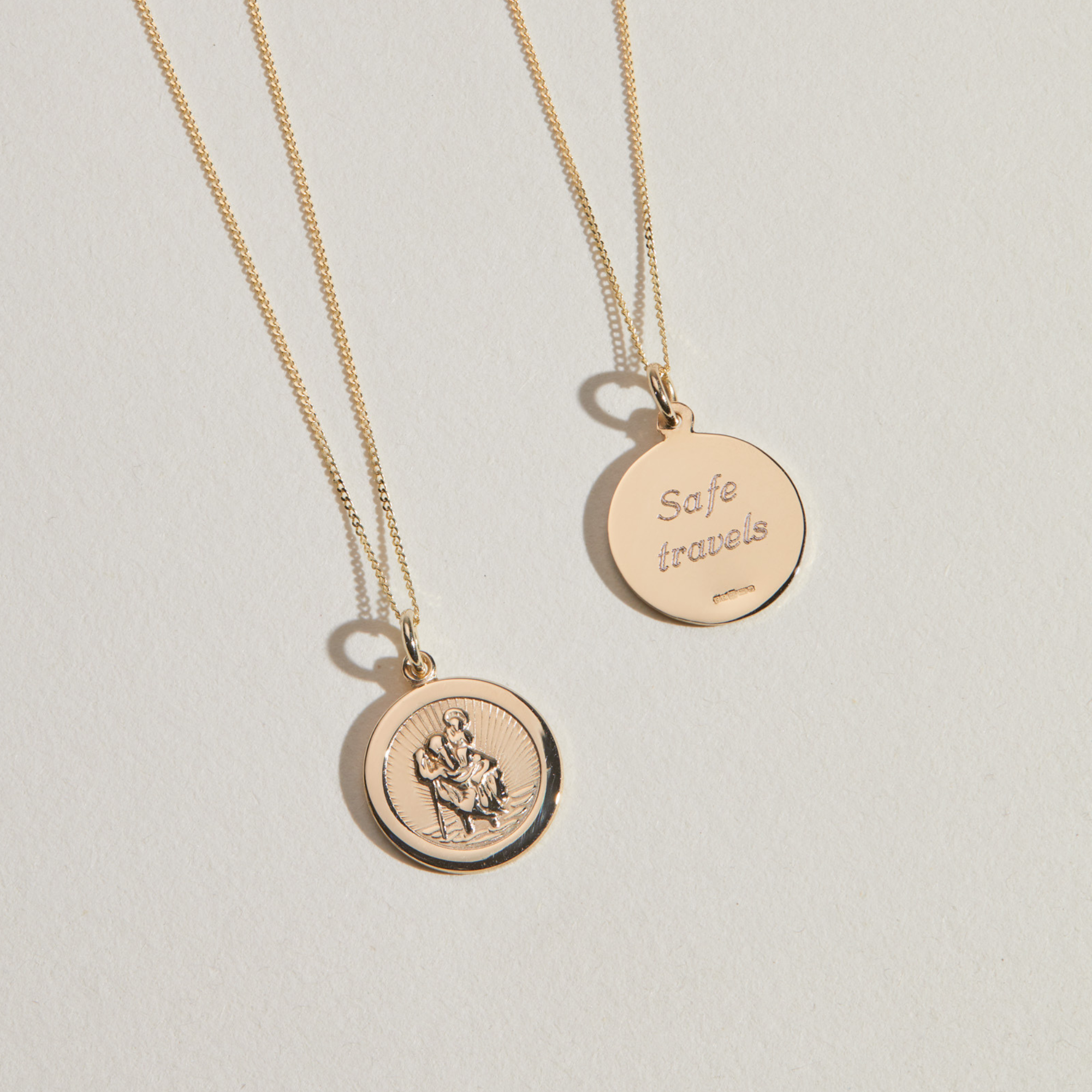 Gold small round St Christopher medallion necklace next to the back of a gold round medallion necklace with the words 'Safe travels' engraved