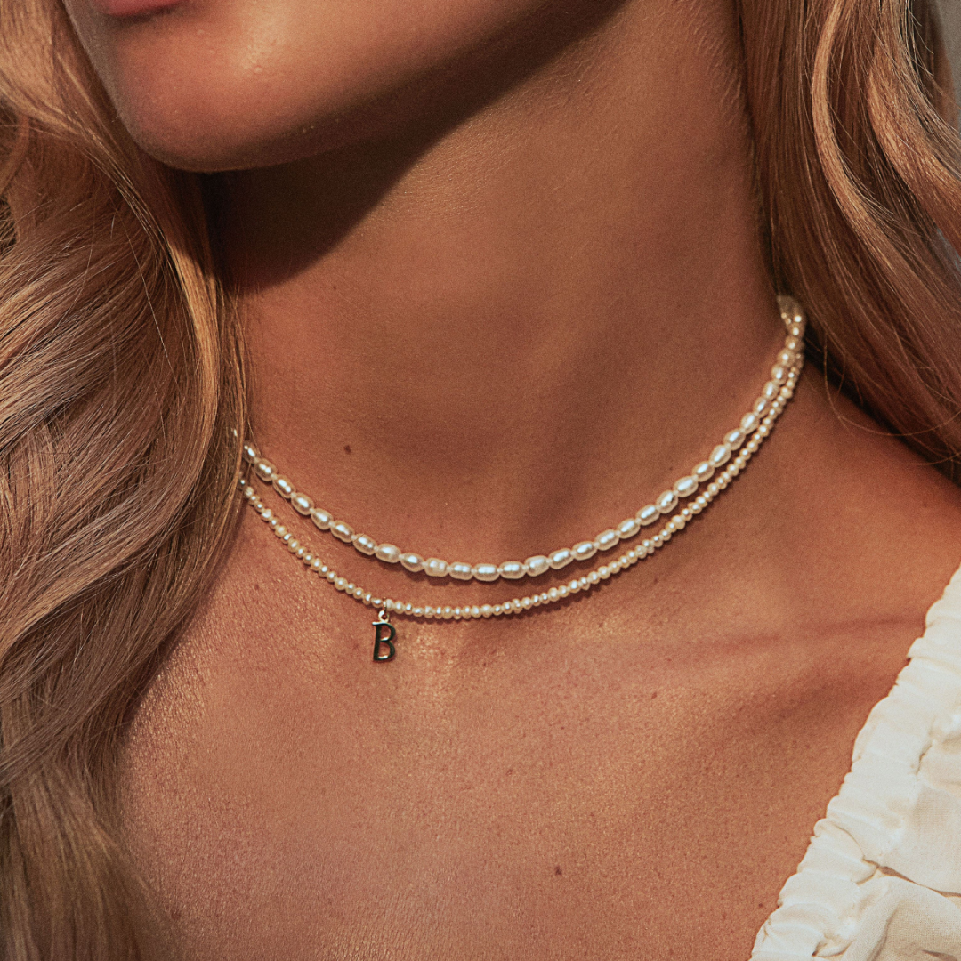 Gold seed pearl choker layered with a gold small pearl initial letter choker letter 'B' around the neck of a blonde woman, close up