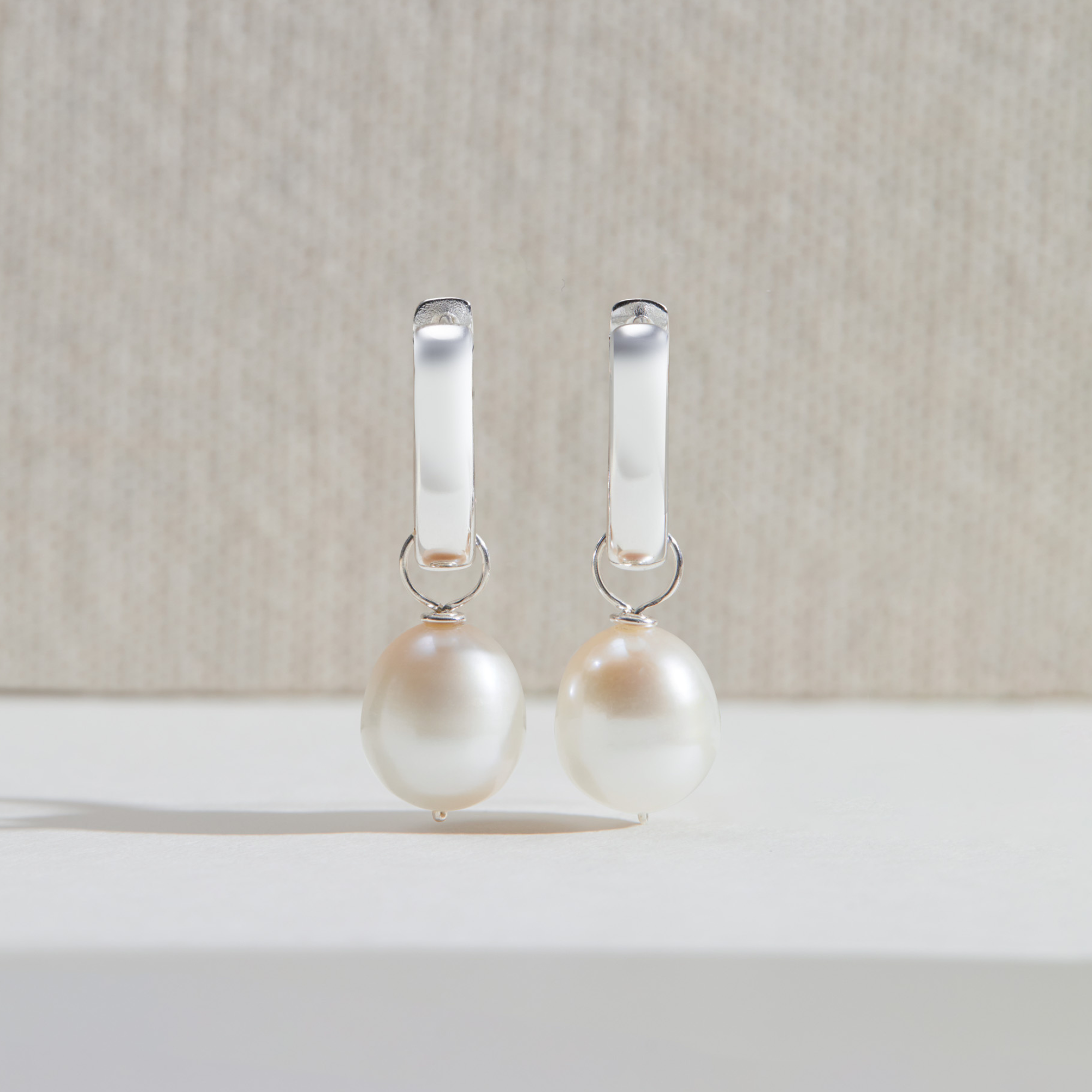 Two silver thick squared hoop pearl drop earrings upright on a white surface with a beige background