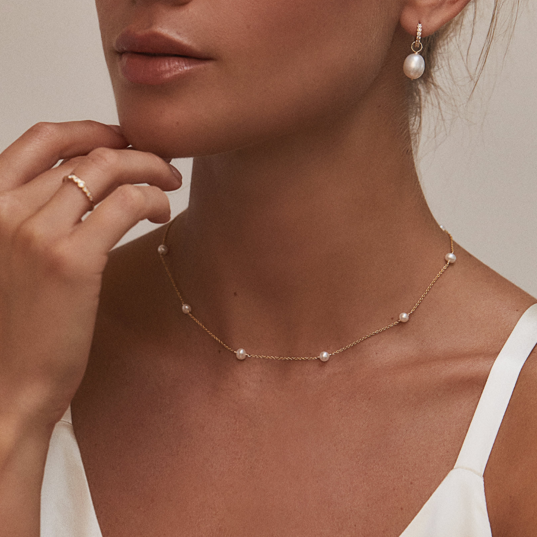 Gold ten pearl choker around a neck paired with a gold huggie pearl drop earring in one ear with her fingers placed under her chin