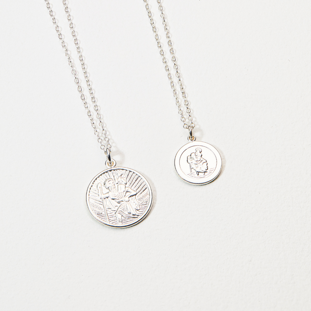 Silver small round St Christopher medallion necklace next to a silver large round St Christopher medallion necklace on a white surface