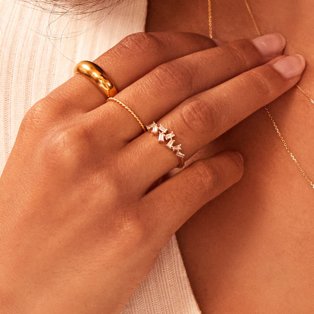 Close up of a hand with a gold thin twisted stacking ring, gold diamond style baguette ring and a gold plain dome ring on the fingers