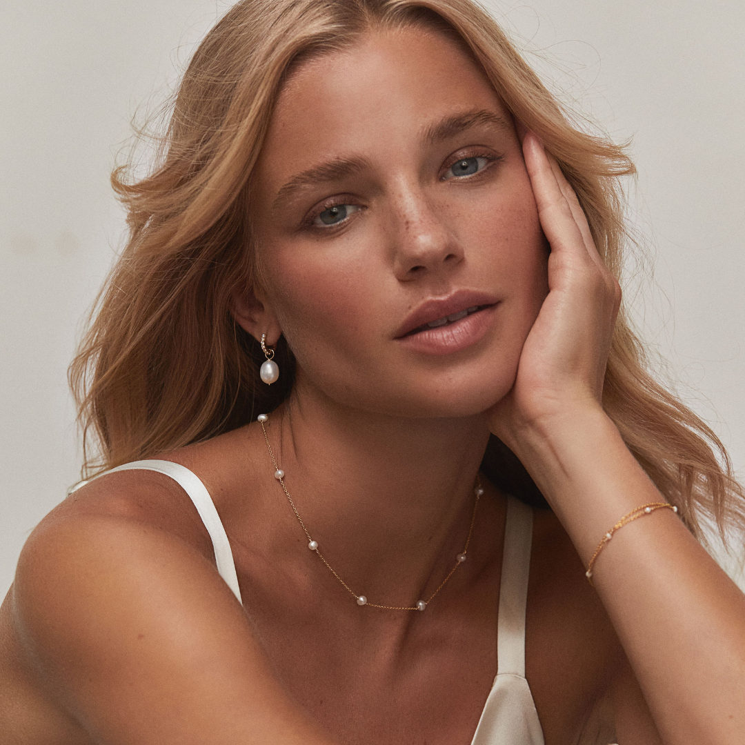 Gold ten pearl choker around the neck of a blonde woman with her hand on her cheek paired with a gold huggie pearl drop earring in one ear and a gold ten pearl bracelet around her wrist