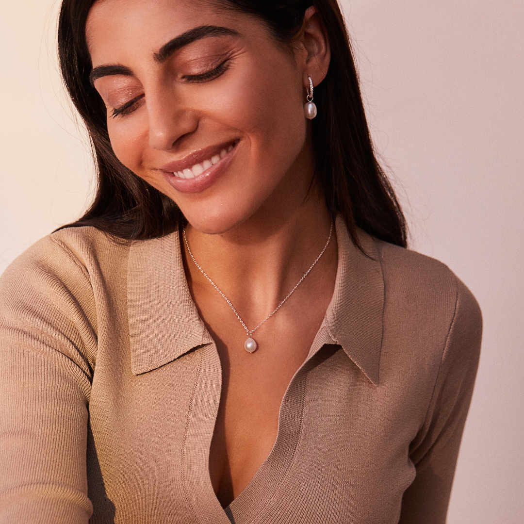 Smiling woman wearing a silver large single pearl necklace and a silver huggie pearl drop earring in one ear lobe 