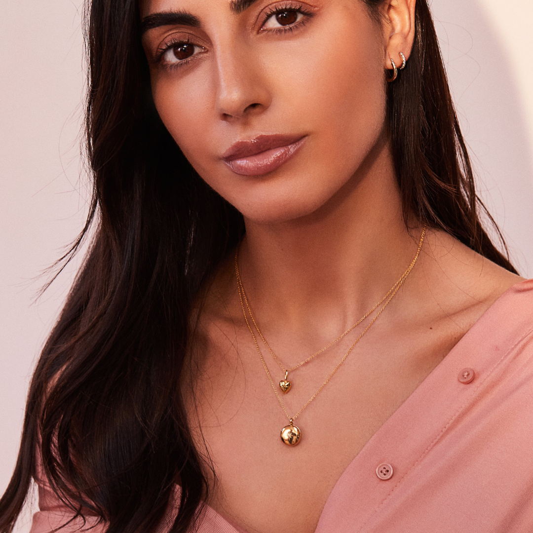 Gold heart pendant necklace around a brunette woman's neck layered with a gold small round diamond locket necklace and wearing a pink shirt