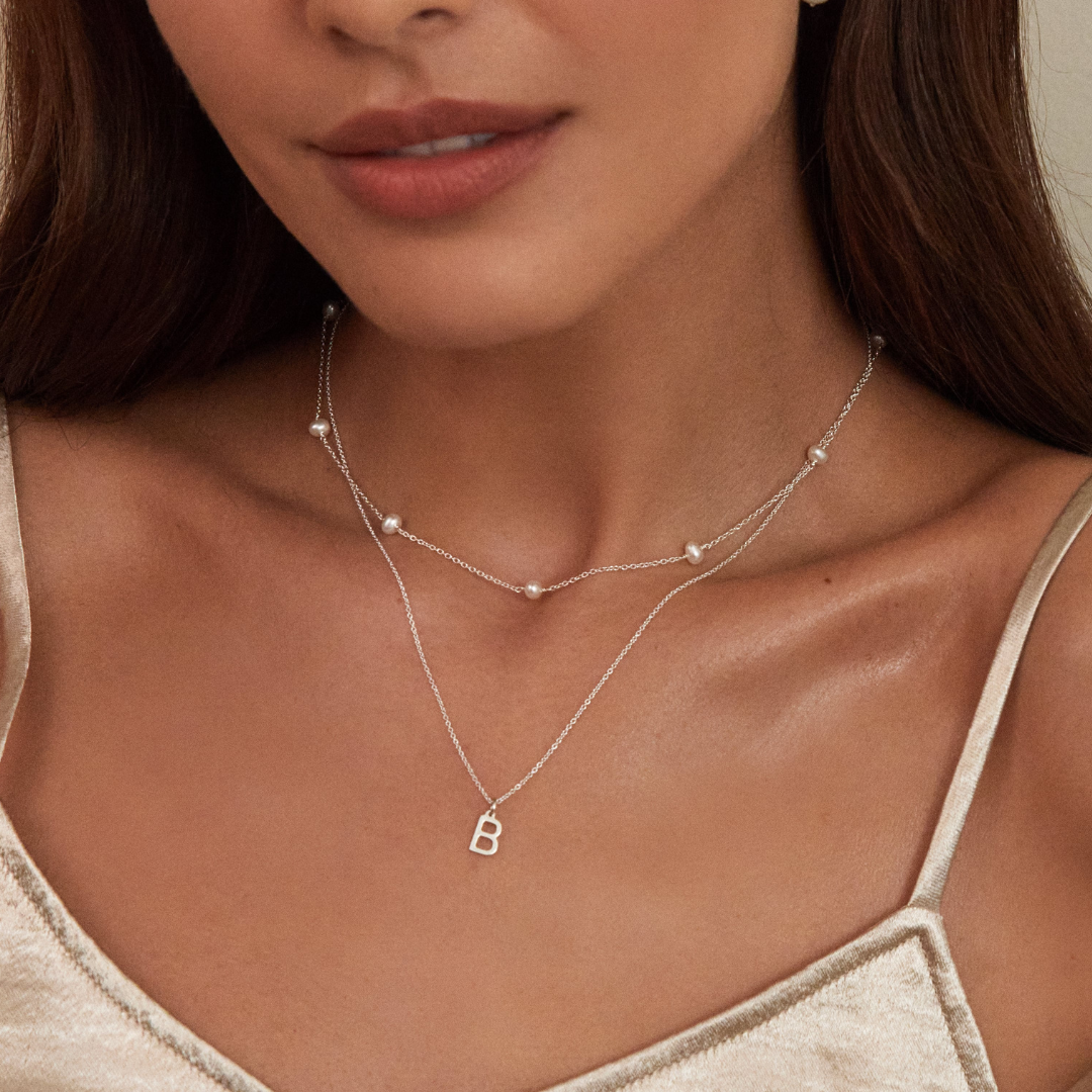 Silver ten pearl choker layered with a silver chain with a silver individual initial charm 'B' around a neck of a brunette woman wearing a gold strappy top