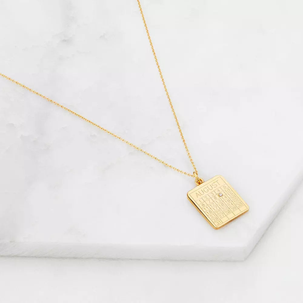 Gold special date calendar necklace 'August' with a gemstone on number '9' on a white marble surface