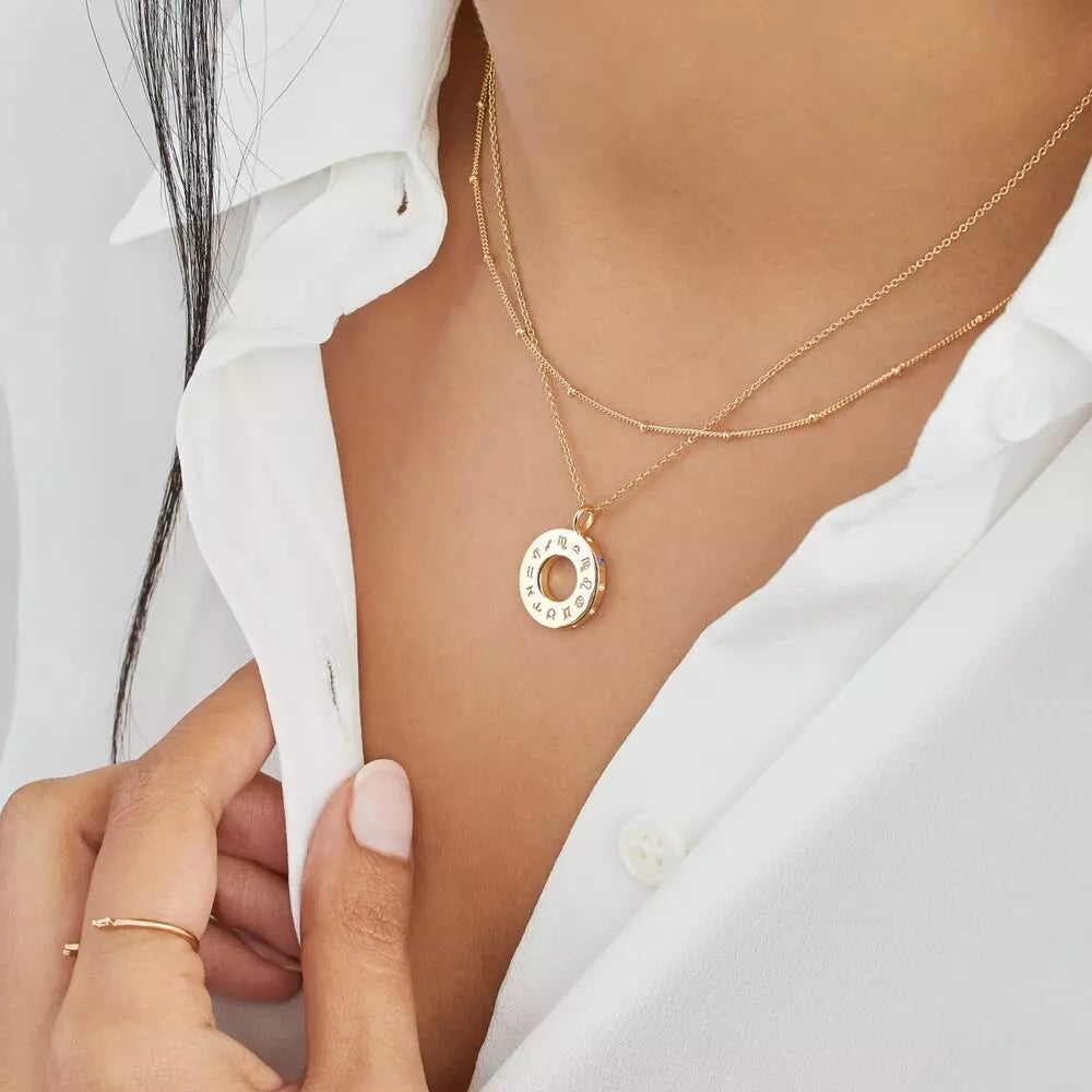 Gold zodiac birthstone necklace layered with a gold satellite chain necklace around the neck of a woman wearing a white shirt