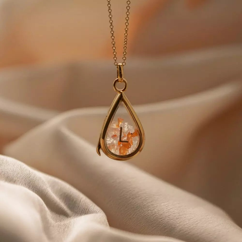 Gold glass gemstone teardrop locket with orange and transparent gemstones in front of beige sheets