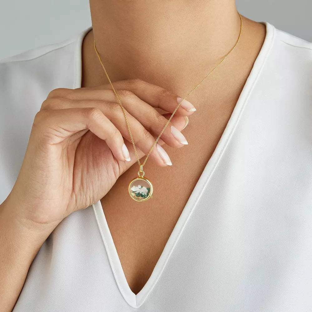 Gold glass gemstone locket with transparent white gemstones and green gemstones around a neck with a white V neck top lifting the necklace off the chest with her hand