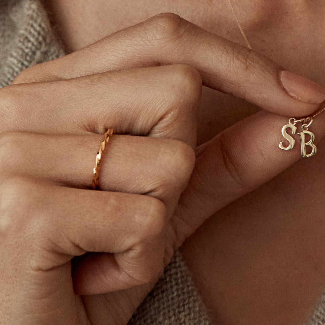 Gold twist ring on a finger of a hand holding up a gold chain necklace with gold cure initial charms 'S' and 'B' on it