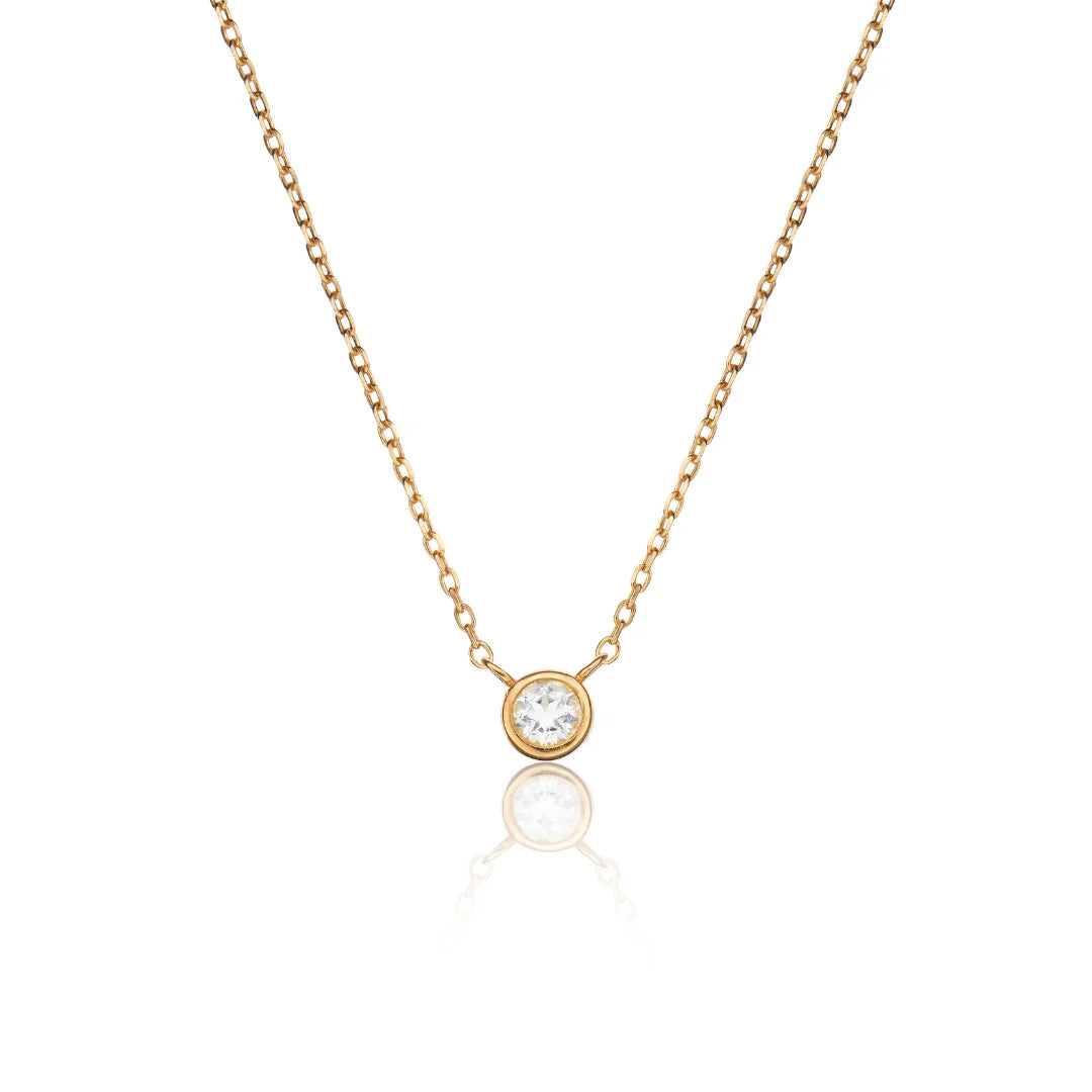 A delicate gold chain necklace with a single round bezel-set clear gemstone pendant, displayed against a clean white background with a soft reflection below.