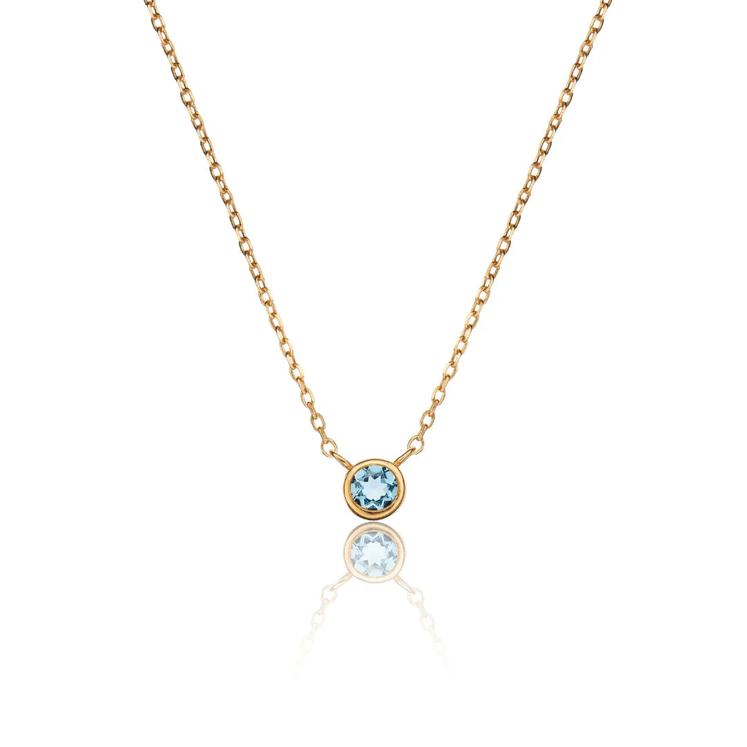 A delicate gold chain necklace featuring a single bezel-set round light blue gemstone pendant, displayed against a clean white background with a soft reflection below.