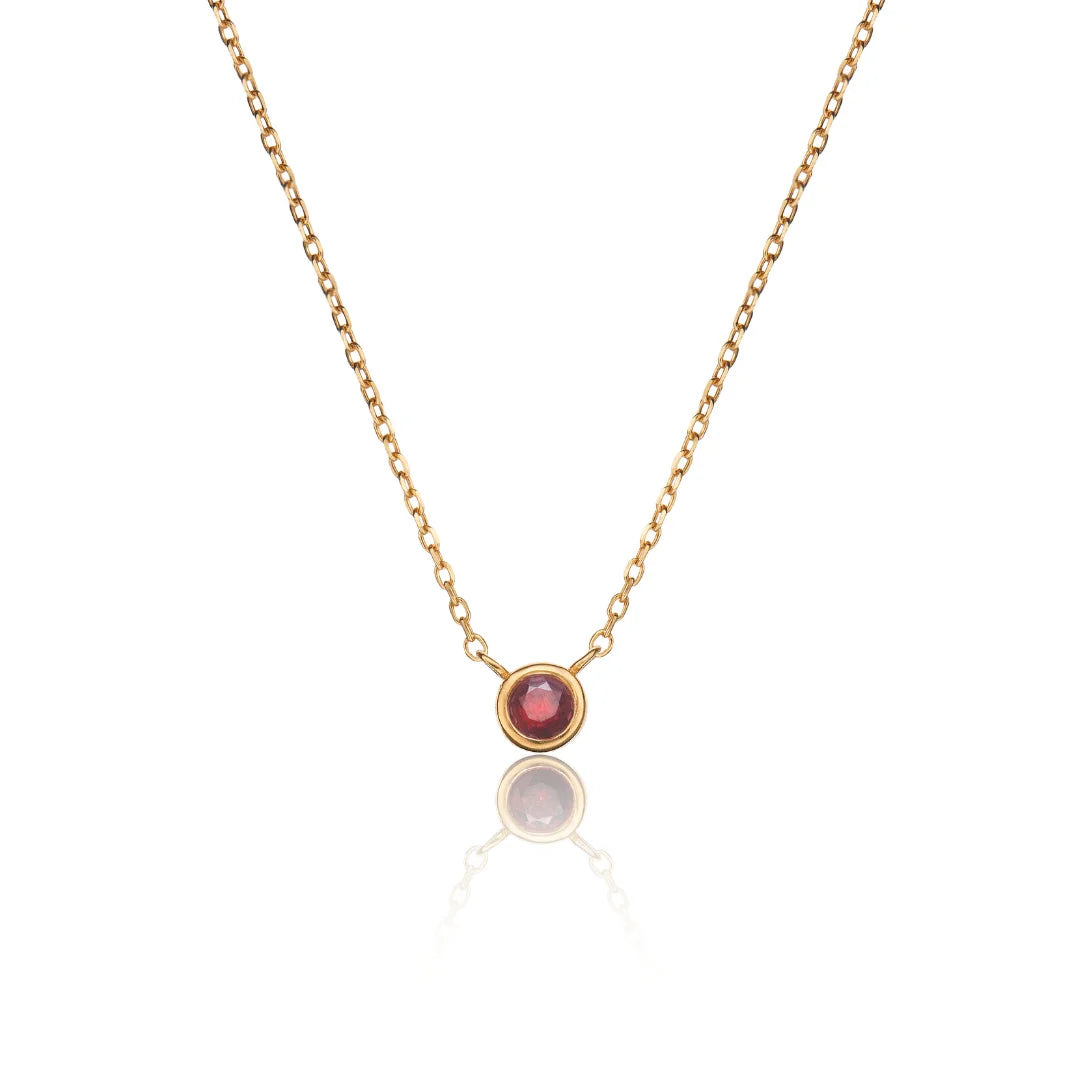 A delicate gold chain necklace featuring a small round bezel-set red gemstone pendant, displayed against a clean white background with a soft reflection below.