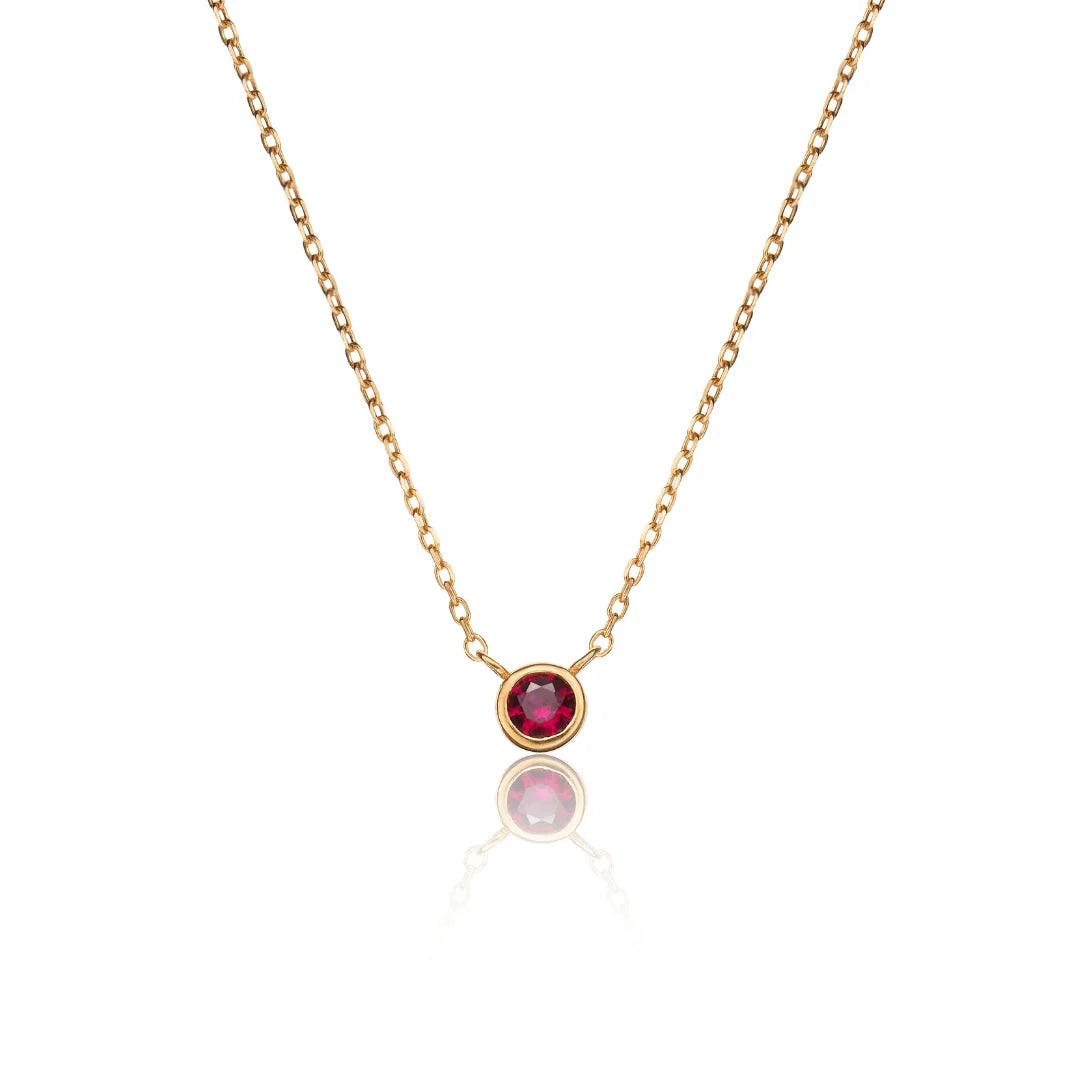 A delicate gold chain necklace featuring a single bezel-set round ruby gemstone pendant, displayed against a clean white background with a soft reflection below.