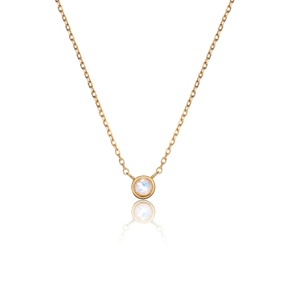 A delicate gold chain necklace featuring a small round bezel-set moonstone pendant, displayed against a clean white background with a soft reflection below.