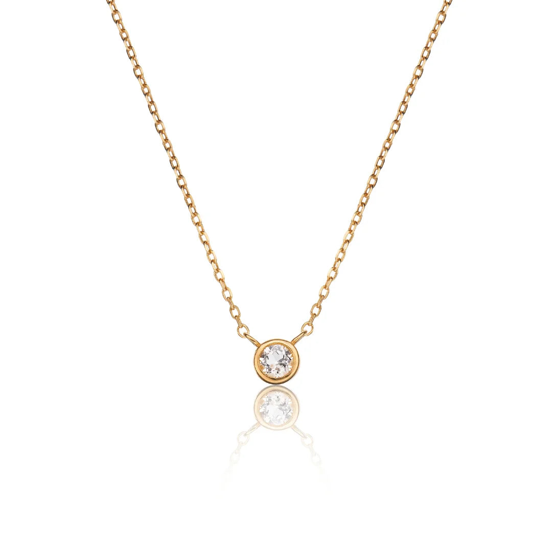 A delicate gold chain necklace with a single bezel-set round clear gemstone pendant, displayed against a clean white background with a soft reflection below.
