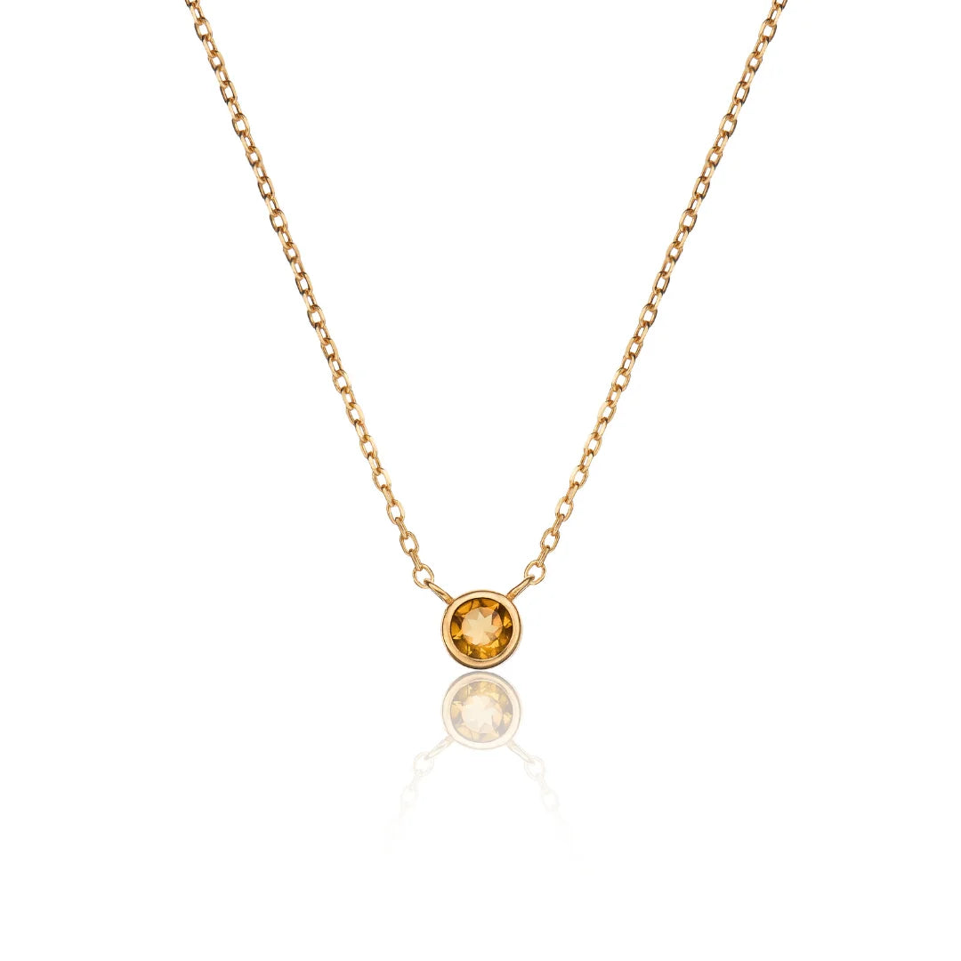 A delicate gold chain necklace with a single bezel-set round orange gemstone pendant, displayed against a clean white background with a soft reflection below.