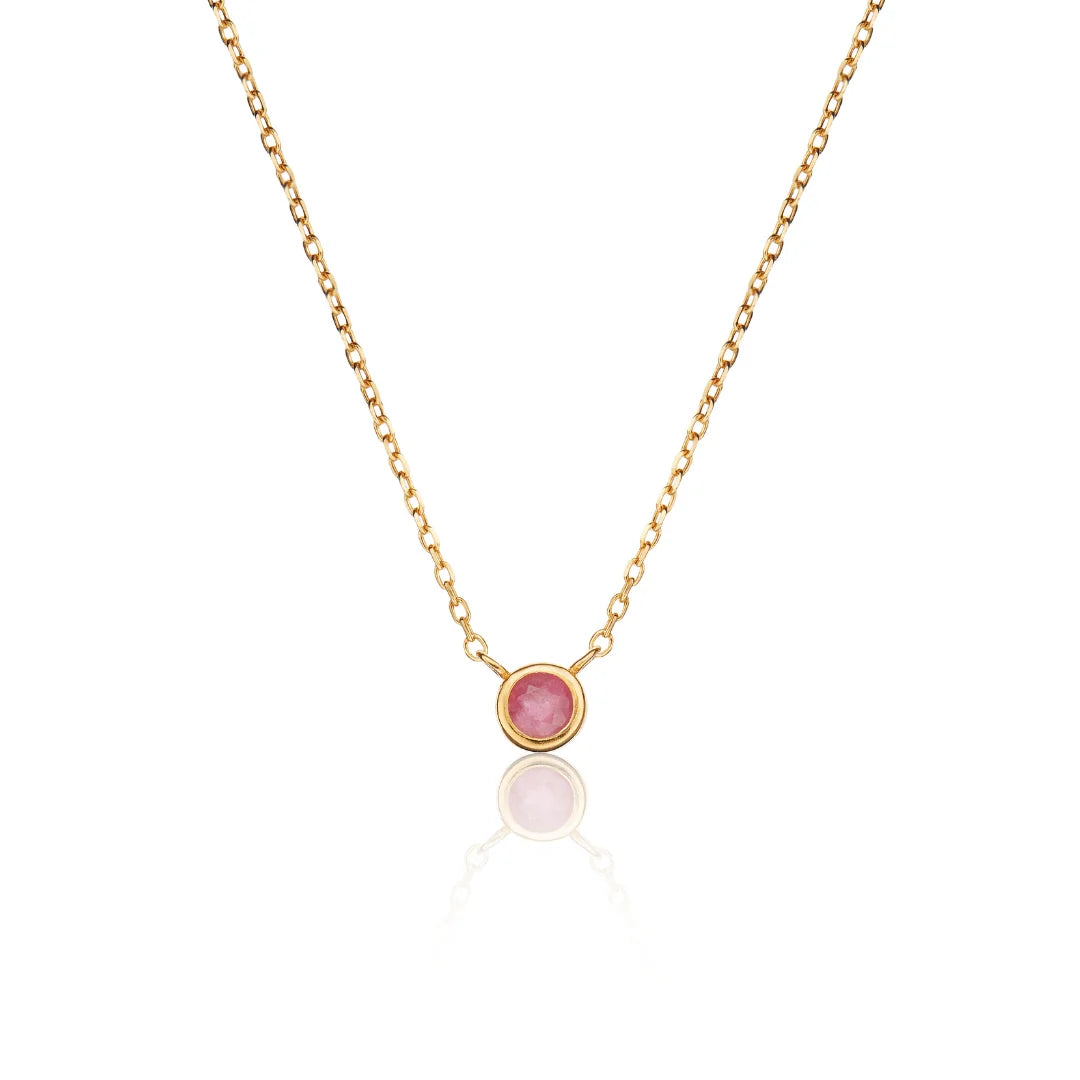 A delicate gold chain necklace featuring a small round bezel-set pink gemstone pendant, displayed against a clean white background with a soft reflection below.