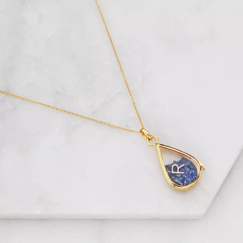 Gold glass gemstone teardrop locket with blue gemstones and a gold R on marble surfaces