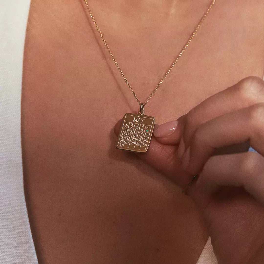 Close up of a gold special date calendar necklace 'May' with a green gemstone on number '10', on the chest of a woman wearing a white top, holding the necklace up with her thumb