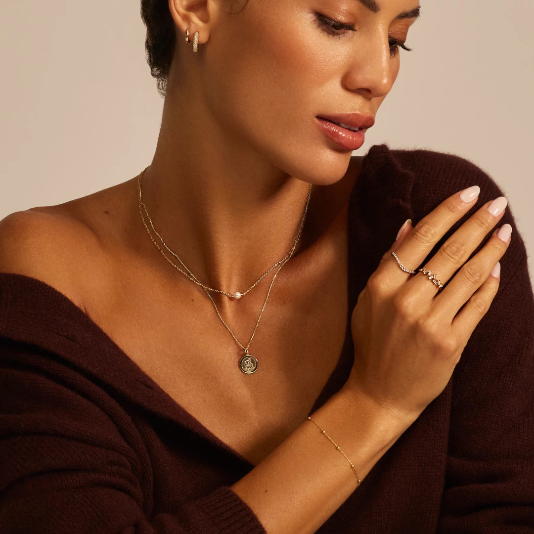 Woman wearing gold jewelry including a necklace, ring, and bracelet against a neutral background