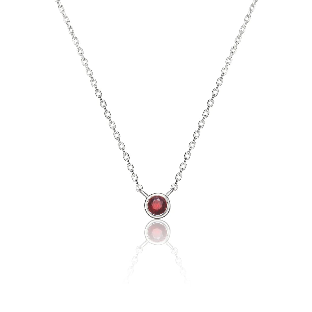A delicate silver chain necklace featuring a single bezel-set round red gemstone pendant, displayed on a clean white background with a soft reflection below.