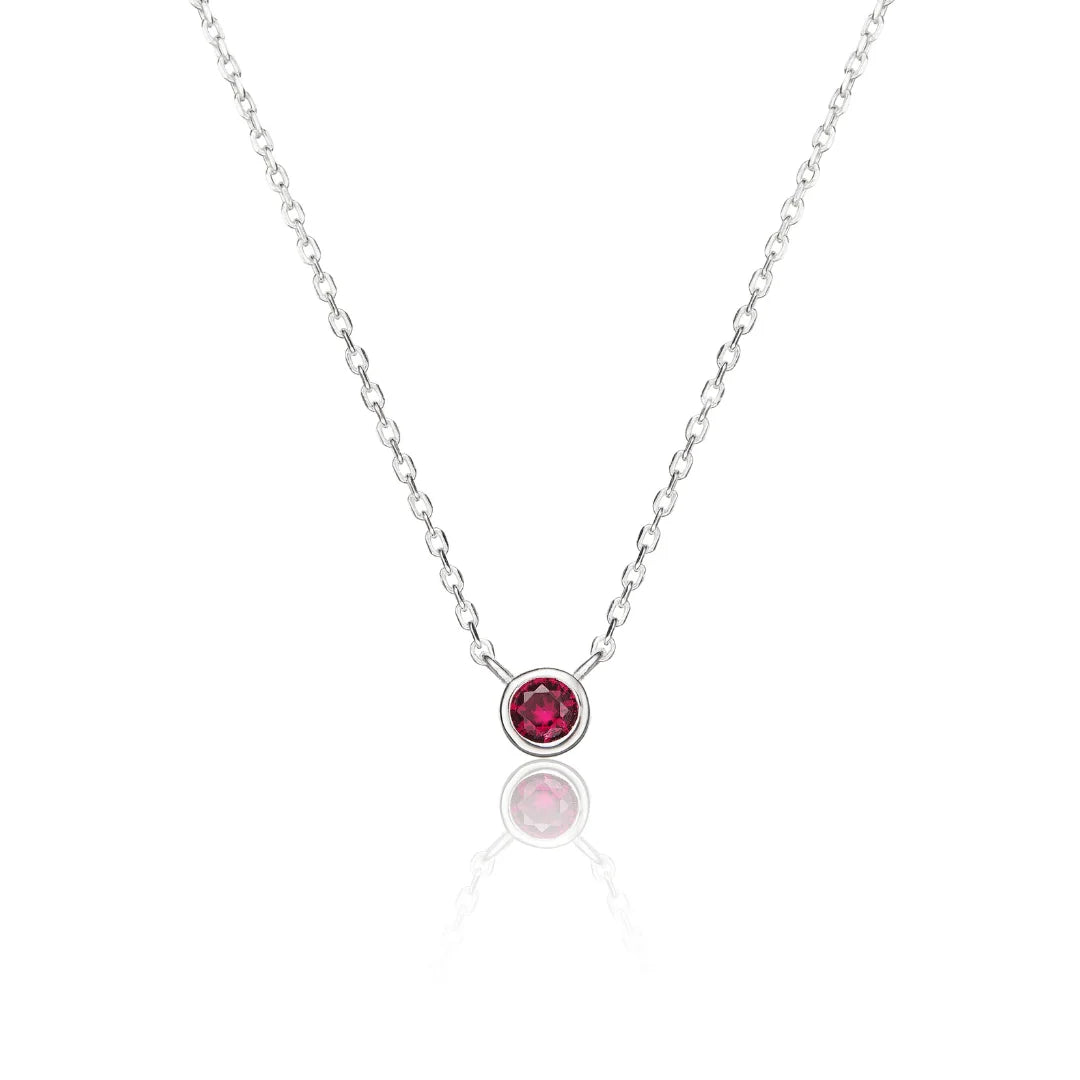 A delicate silver chain necklace with a small round bezel-set red ruby gemstone pendant, displayed on a clean white background with a soft reflection below.