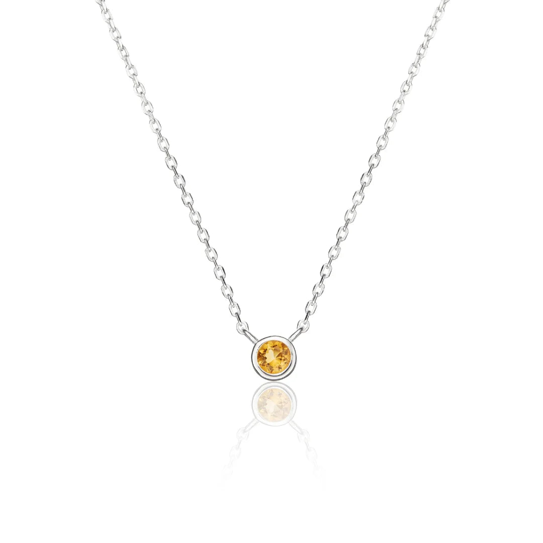 A delicate silver chain necklace featuring a single round bezel-set yellow gemstone pendant, displayed on a clean white background with a soft reflection below.