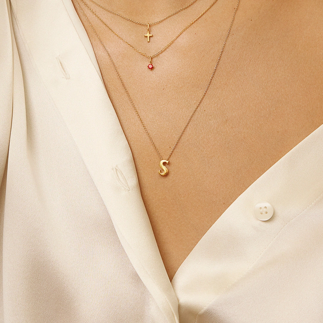 Close-up of a person wearing layered gold necklaces over a cream satin shirt. The necklaces include a small cross pendant, a tiny red gemstone pendant, and a gold letter 'S' pendant.