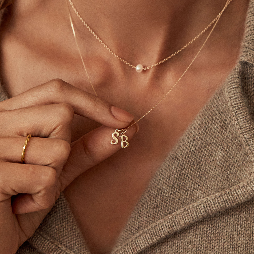 Solid gold curve initial letter necklace with the initials 'S' and 'B' around the neck of woman holding it up with her finger tips. She is also wearing a gold single pearl choker around her neck and a gold thin twisted stacking ring on her finger 